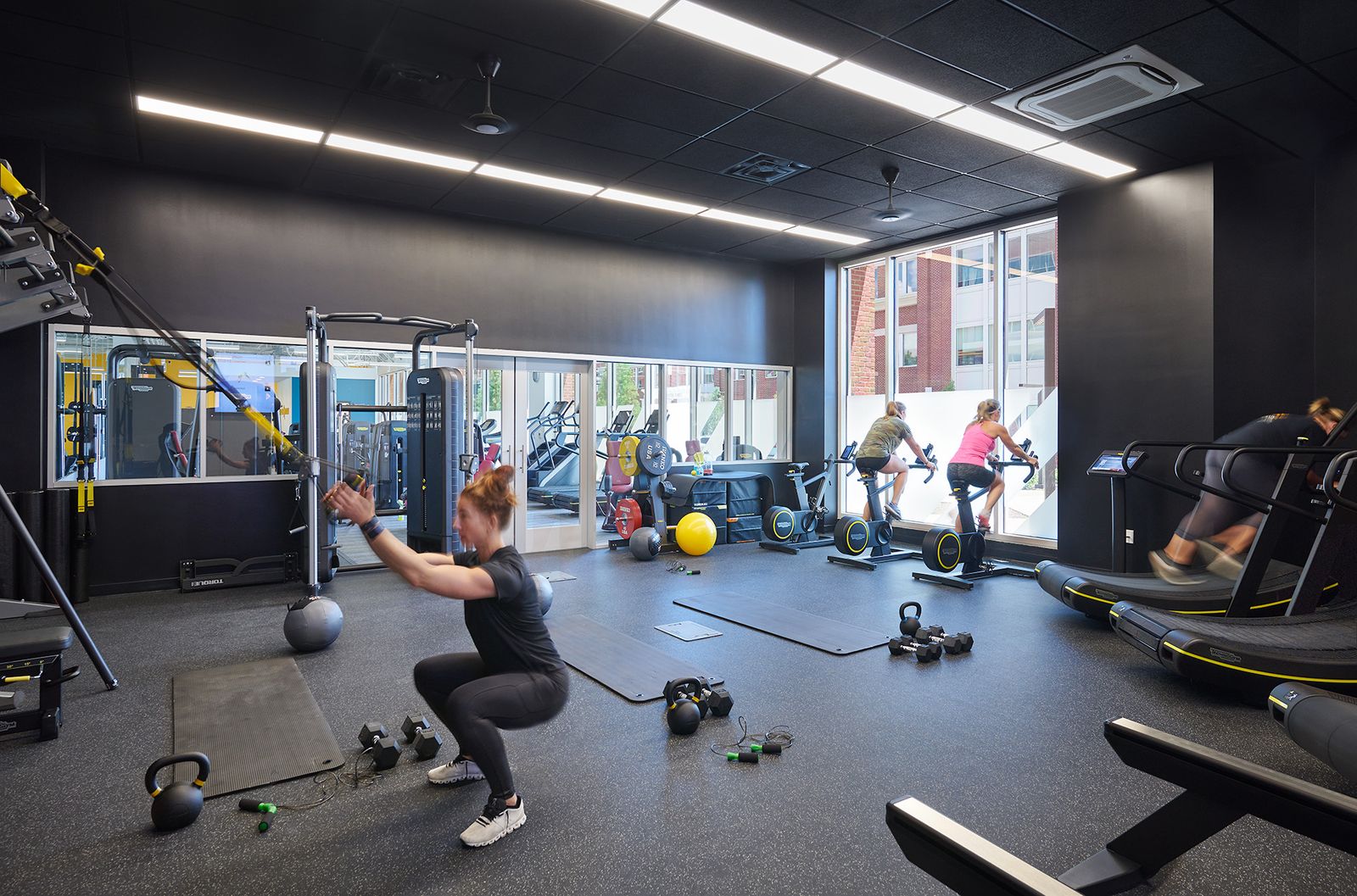 A member squats on a suspension trainer while others ride bikes by the windows in a wide training studio