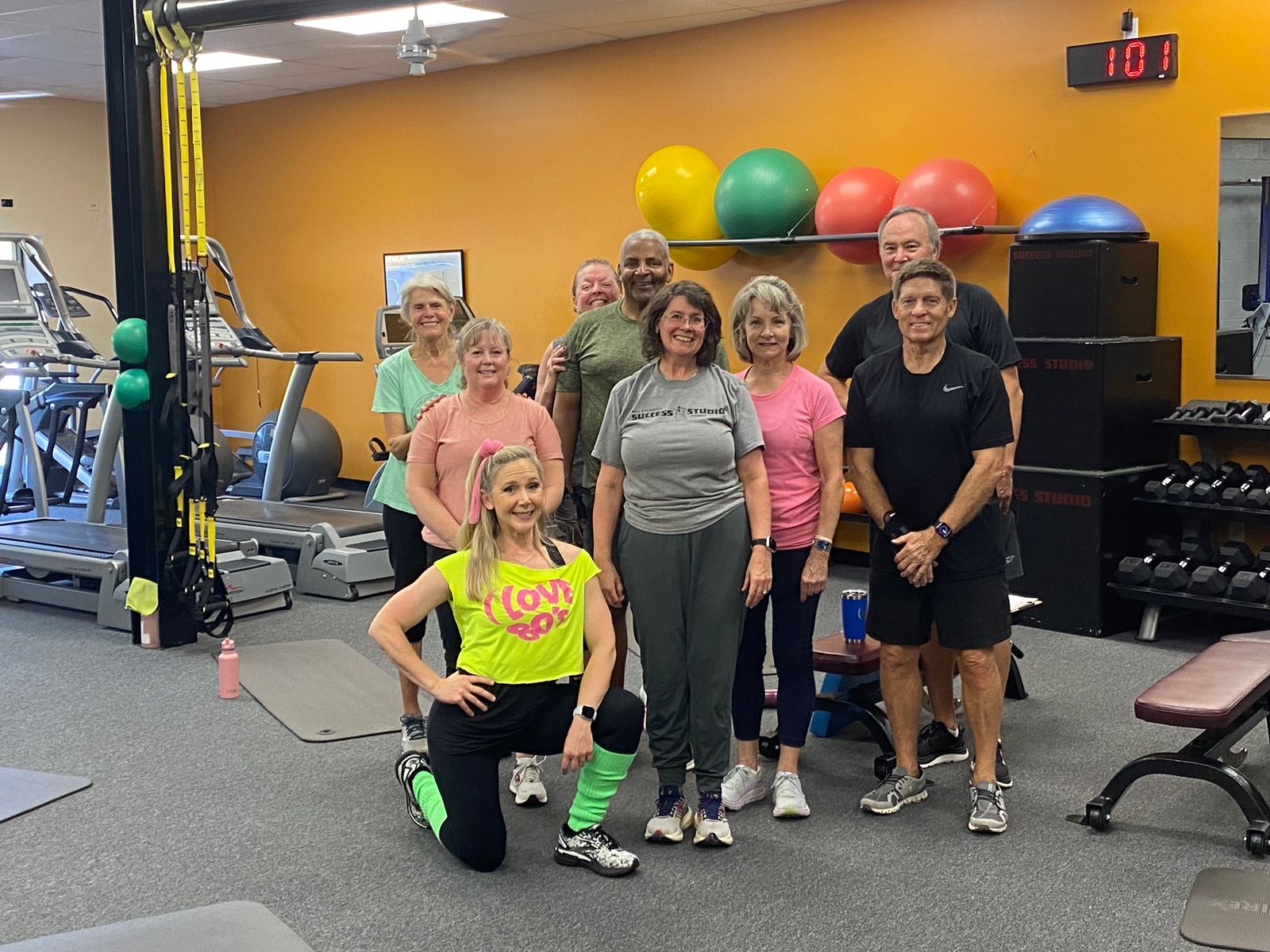 Eleven members pose after a group class, their instructor kneeling in front in an ’80s costume