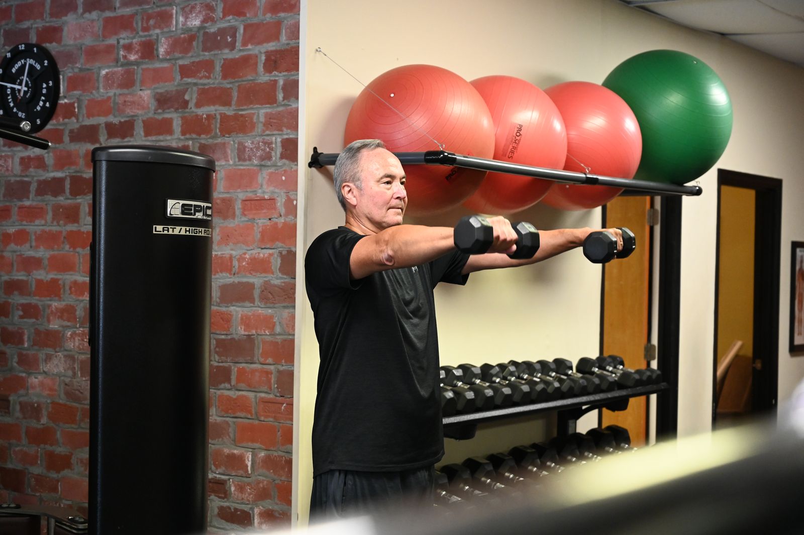 A grey-haired member raises a pair of dumbbells in front of him beside a brick wall, with exercise balls racked behind.