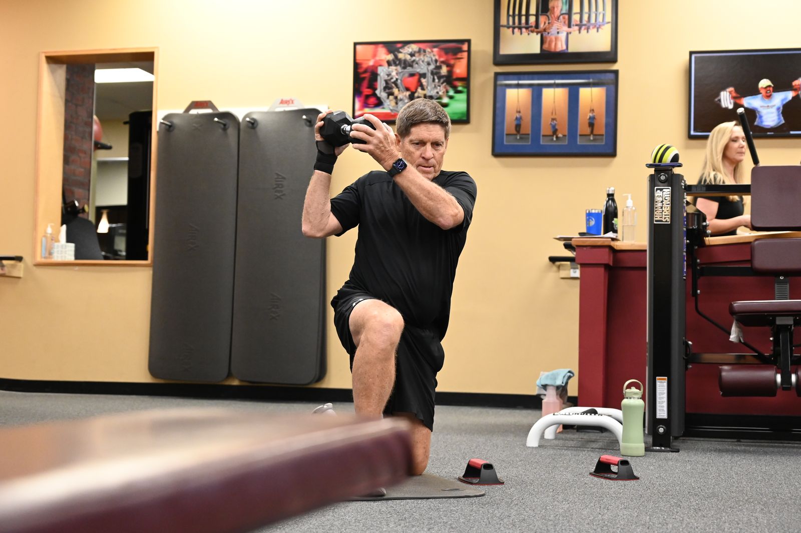 A member in a half-kneeling stance presses a dumbbell overhead near the Ivy Road front desk