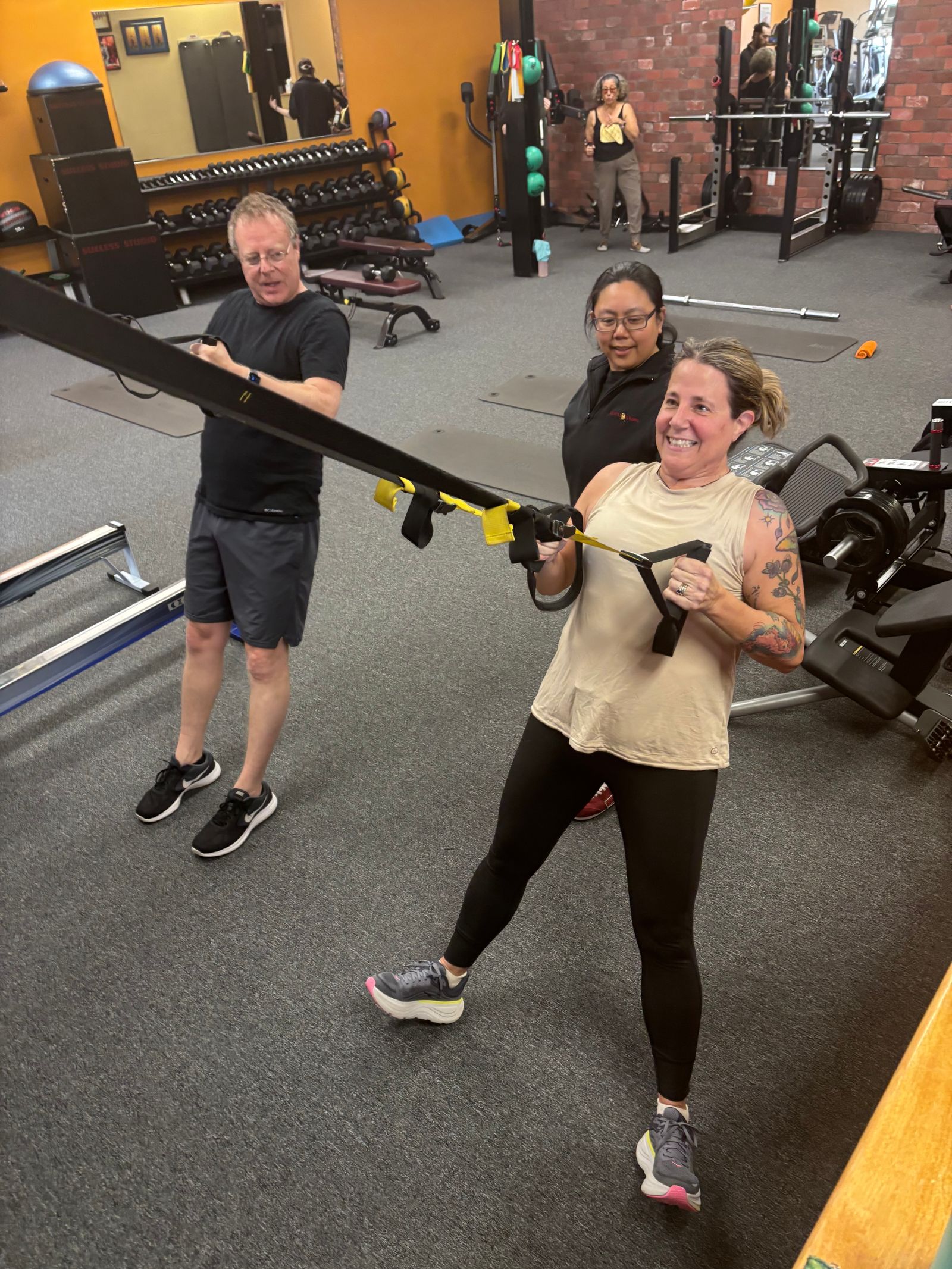 A trainer stands behind a member doing a suspension-trainer row while another member works the next strap