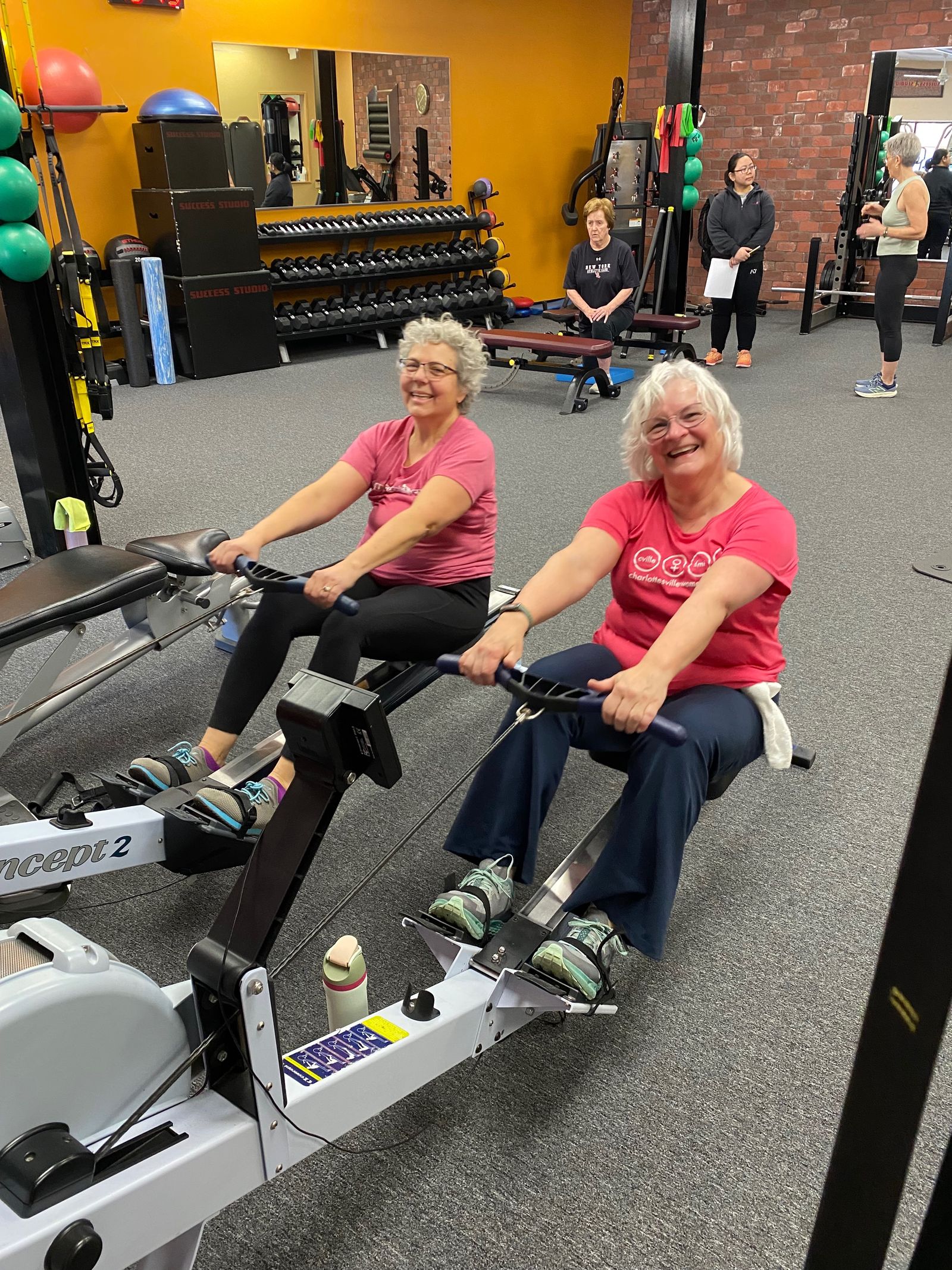 Two women laugh side by side on rowing machines.