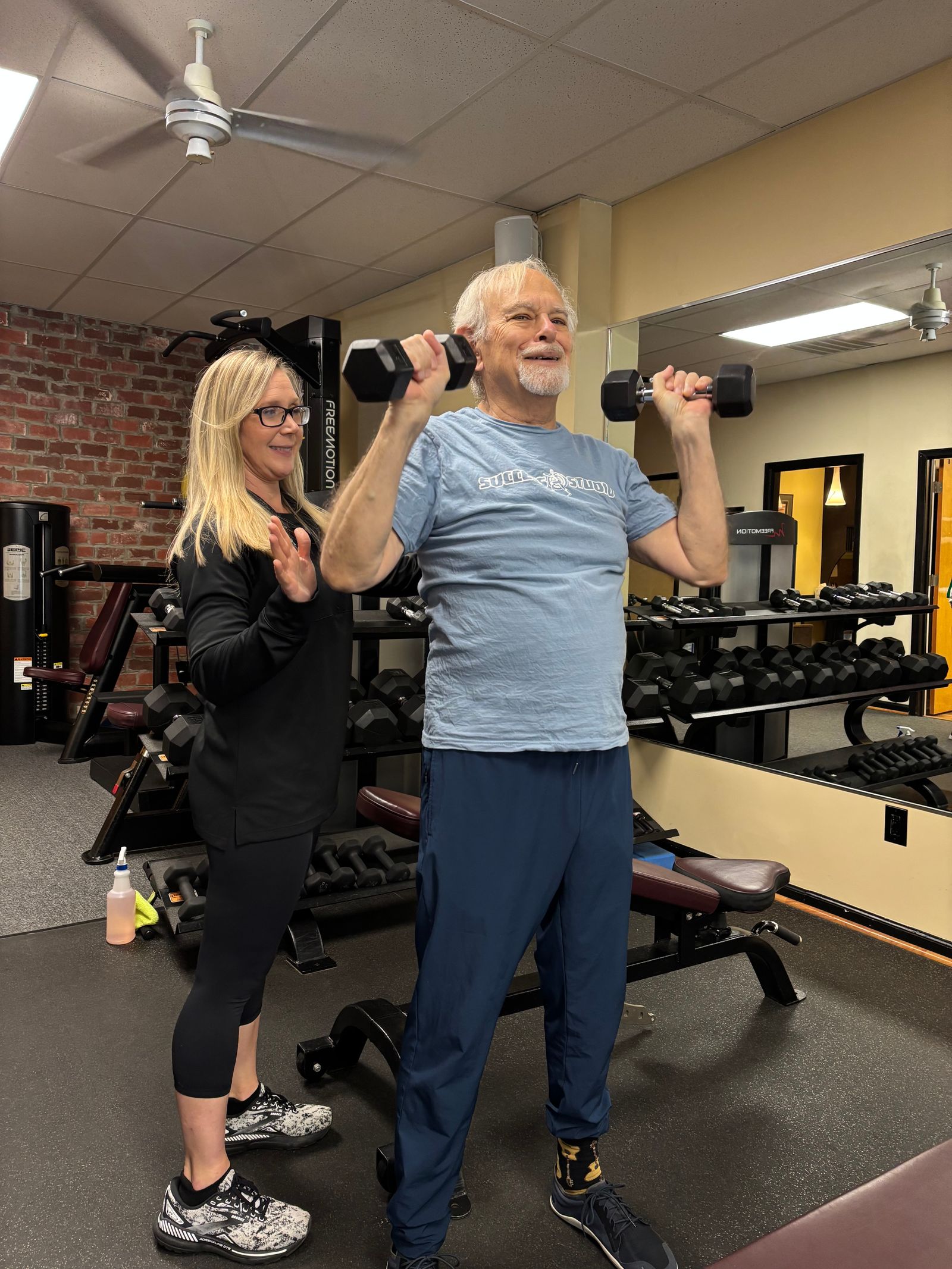 A trainer guides a member through standing dumbbell curls beside the dumbbell rack