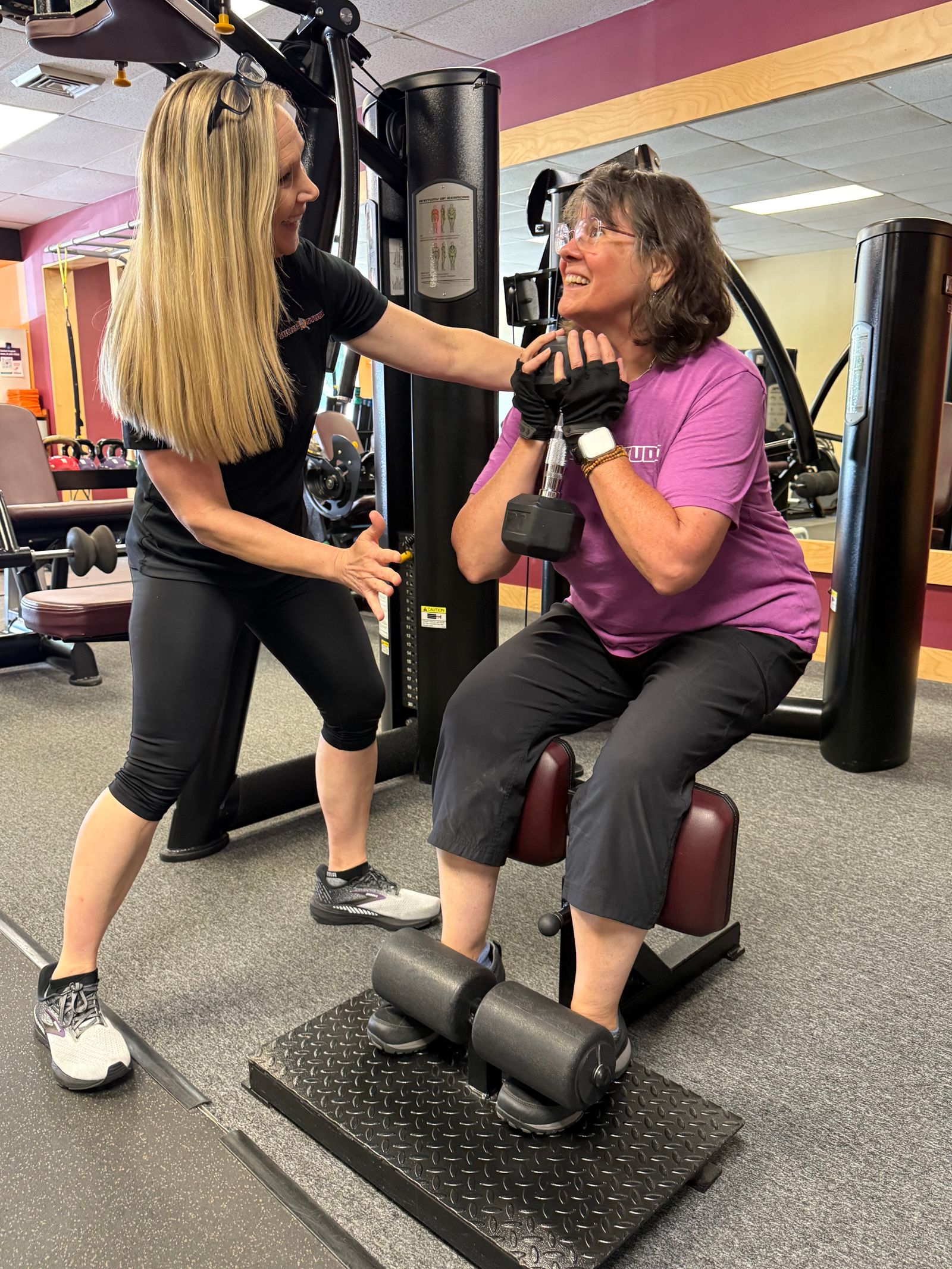 A trainer leans in, training a smiling member through a seated dumbbell exercise.