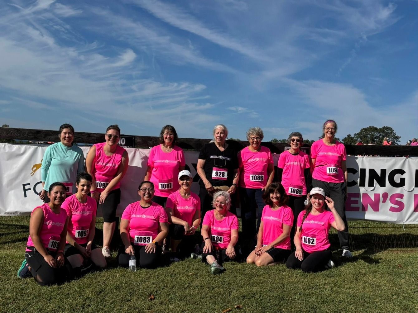 Fourteen women in pink Success Studio race shirts and bibs pose in front of a race banner