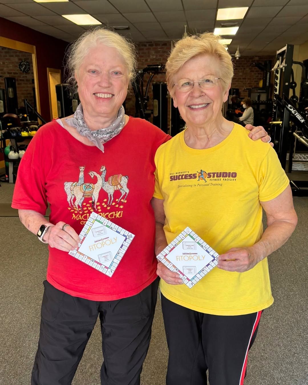 Two members smile arm in arm, each holding a game card from a studio challenge