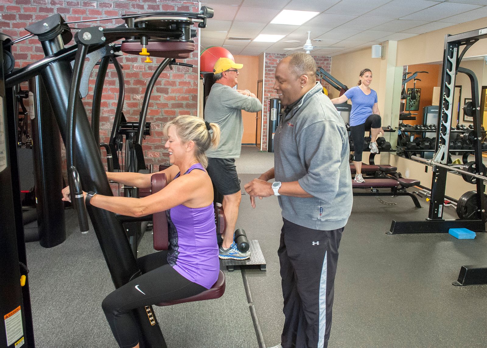 A trainer training a smiling member through a chest press on a machine while others work out behind them