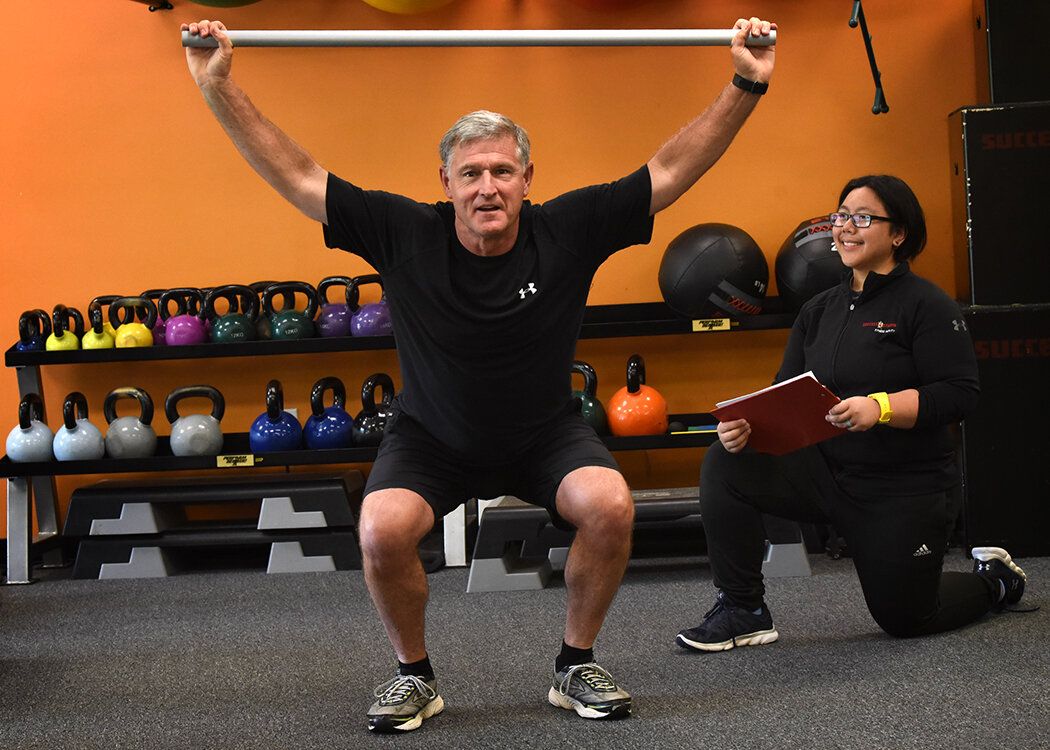 A member holds a dowel overhead in a squat while a trainer kneels beside him taking notes