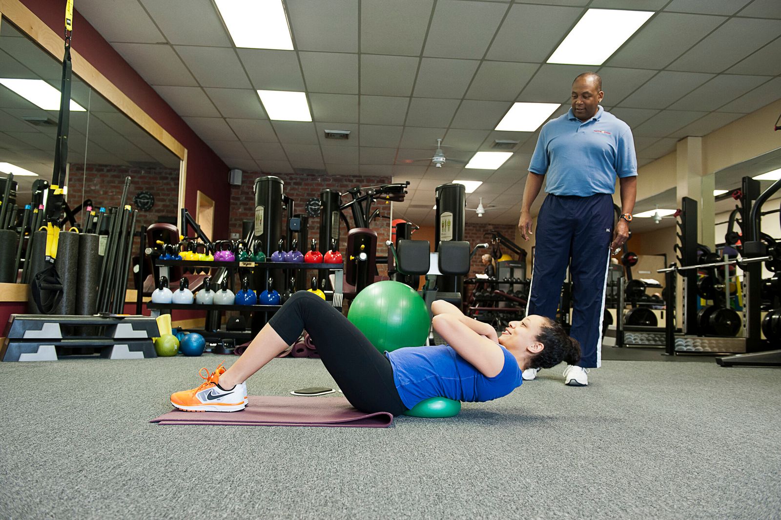 A trainer watches a member do a crunch over a stability ball in the wide training studio
